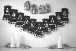 Ten cheerleader uniforms, each adorned with a letter, are hung on a wall in a vee shape to spell out DISABILITY. Pom-poms and megaphones complete the tableau.
