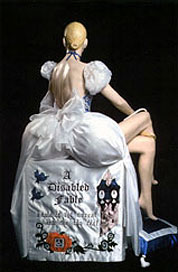 A life size Cinderella manequin, complete with fairytale ballgown, is seated atop an ornate banner that reads A Disabled Fable. There is a blue and white pillow on the floor in the background.