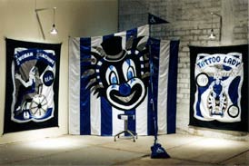 This installation features a pennant two large appliqued circus-style banners (one featuring a woman in a wheelchair and one promising a Tattoo Lady), a blue and white striped stool and a blue and white striped fabric chamber adorned with a giant clown face.