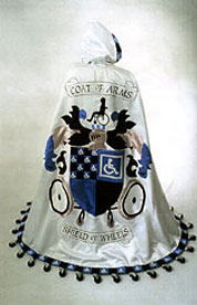 A white hooded cloak is emblazoned with Coat of Arms and appliqued with a woman in a wheelchair in profile silhouette and a crest featuring the Wheelchair Symbol of Access. The hem of the cloak is fringed with casters (small wheels).