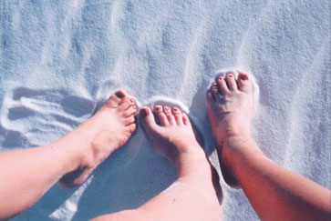 three human calves and feet (two right and one left) with with toes displaying polished toenails make imprints in rippling sand.