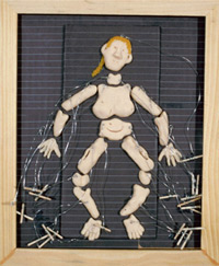 This image appears to be a mixed media collage, with a blonde wood frame, which is about two thirds as wide as tall. Inside the frame, is a background of black fabric horizontally striped with grey. Against this background is a slightly raised, centered, veritcal rectangle half the width and about seven eights of length of the piece in size. This too is covered in black fabric with grey horizontal stripes. Upon this bed, is a disjointed puppet figure of a woman, lying on her back, with arms splayed and crooked at either side, legs spread at knees, with ankles coming back in toward each other. The puppet figure has separate pieces for head, neck, chest, pelvis, upper arms, forearms, hands, thighs, knees, calves and feet. These individual pieces have holes with varying lengths transparent tubing attached and terminating in the wood  sticks used by a puppeteer.